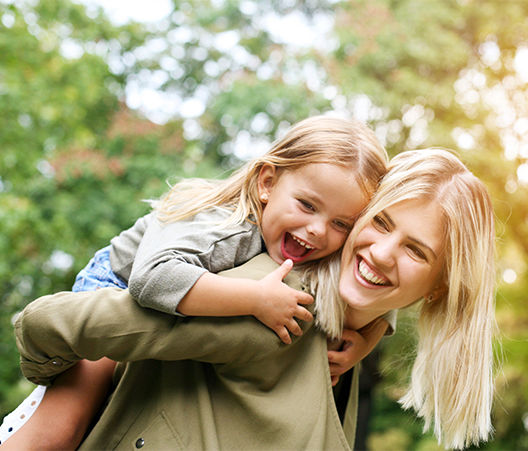 Mother giving her daughter a piggy back ride outside while they play in the bright sun